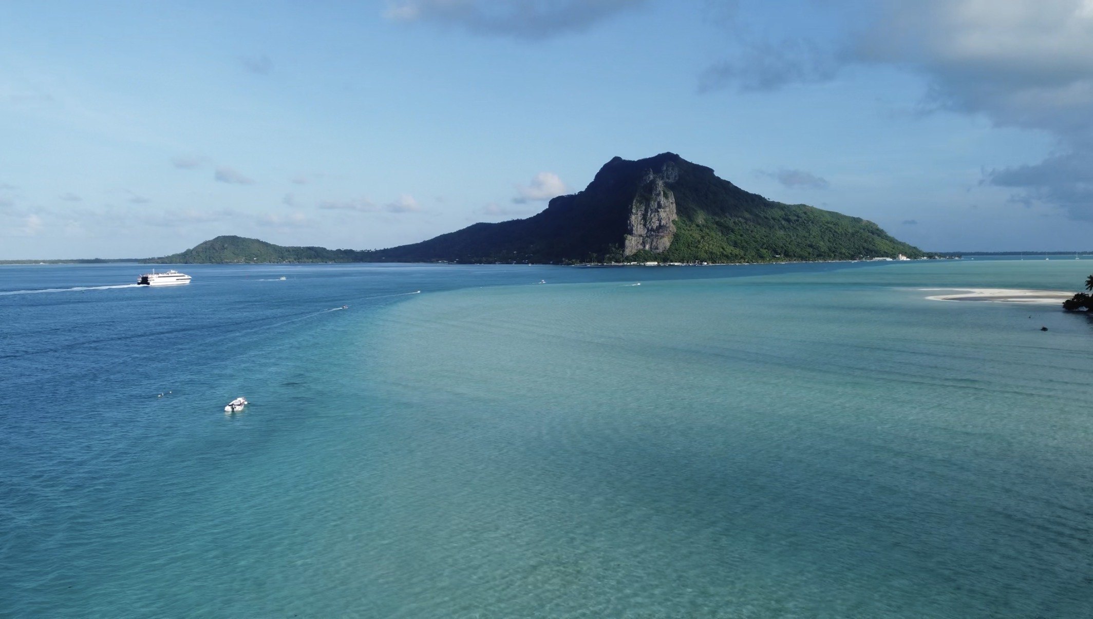 An island rising steeply across a turquoise lagoon, a ferry and a
              small boat out on the water