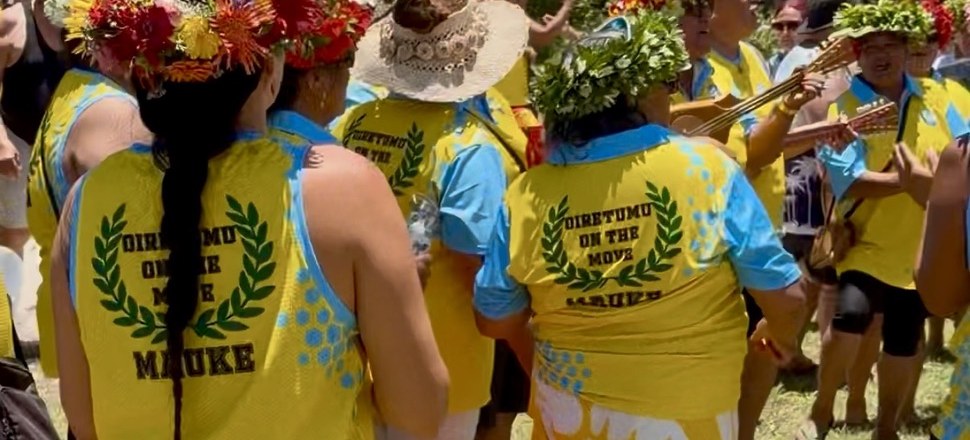 The people of Oiretumu, Maʻuke, in yellow shirts reading
              &lsquo;Oiretumu on the Move&rsquo;, wearing ʻei katu of
              flowers and leaves, with ʻukulele playing among them